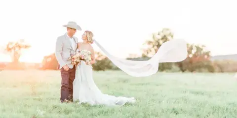 A bride and groom standing in a grassy field at sunset, with the bride's veil blowing in the wind.