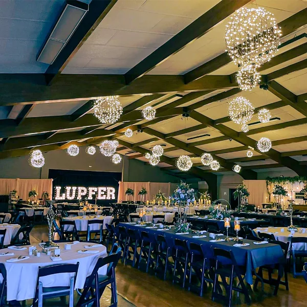 Interior of a large event hall with a wooden beam ceiling, set up with round tables and hanging spherical lights for a wedding reception.