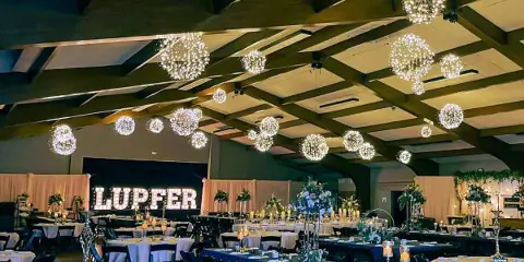 Interior of a large event hall with a wooden beam ceiling, set up with round tables and hanging spherical lights for a wedding reception.