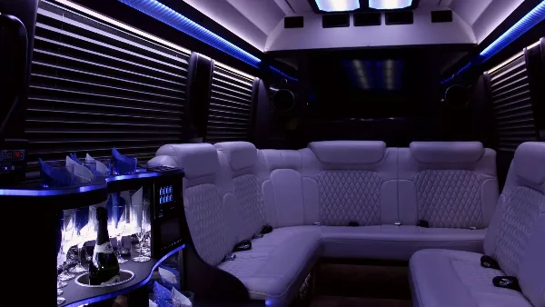 Luxury limousine interior featuring white leather seating, ambient blue lighting, and a stocked bar.