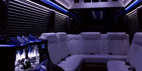 Luxury limousine interior featuring white leather seating, ambient blue lighting, and a stocked bar.