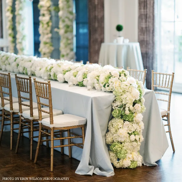 A long banquet table draped in white linen with a lush, cascading floral centerpiece of white roses and hydrangeas, surrounded by gold chiavari chairs.