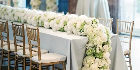 A long banquet table draped in white linen with a lush, cascading floral centerpiece of white roses and hydrangeas, surrounded by gold chiavari chairs.