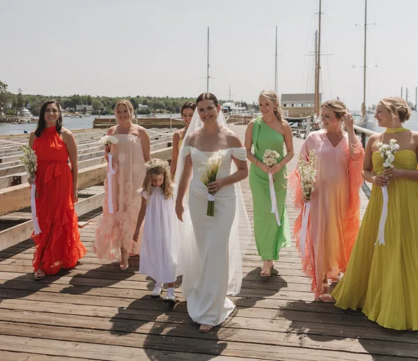 Bride and bridesmaids in colorful dresses walking on a wooden dock at a coastal harbor wedding.