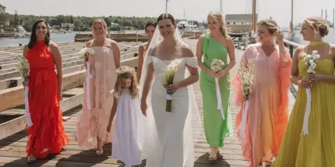Bride and bridesmaids in colorful dresses walking on a wooden dock at a coastal harbor wedding.