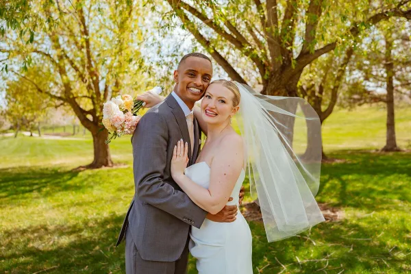 A smiling bride and groom posing together outdoors under a tree, with the bride holding a bouquet and wearing a veil.