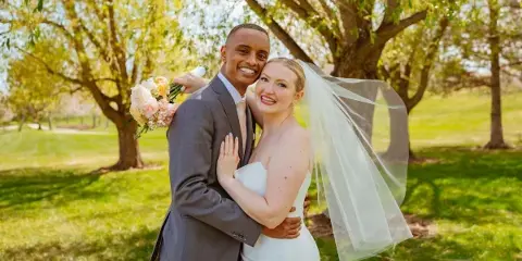A smiling bride and groom posing together outdoors under a tree, with the bride holding a bouquet and wearing a veil.