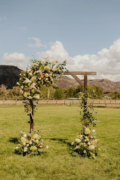 A wooden wedding arch decorated with flowers and greenery set in a grassy field with mountains in the background.