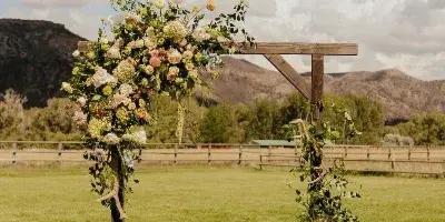 A wooden wedding arch decorated with flowers and greenery set in a grassy field with mountains in the background.