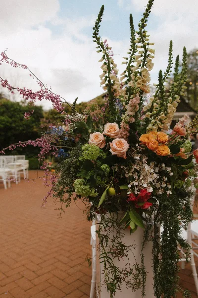 A tall, organic wedding floral arrangement featuring cream roses, foxgloves, and cascading greenery on a white pedestal.