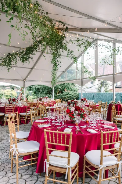A wedding reception setup inside a tent with red tablecloths, gold chiavari chairs, floral centerpieces, and hanging greenery.