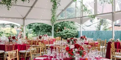 A wedding reception setup inside a tent with red tablecloths, gold chiavari chairs, floral centerpieces, and hanging greenery.