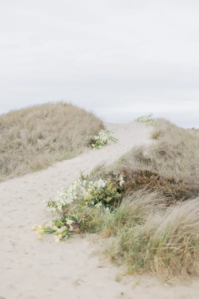 A wedding bouquet with white and yellow flowers resting on a sandy path between dune grass.