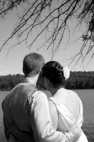 Black and white photograph of a bride and groom embracing from behind, standing by a lake with trees in the background.
