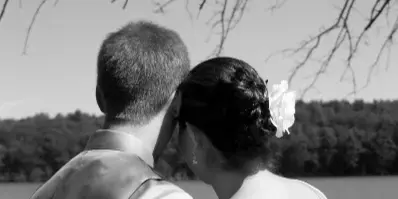 Black and white photograph of a bride and groom embracing from behind, standing by a lake with trees in the background.