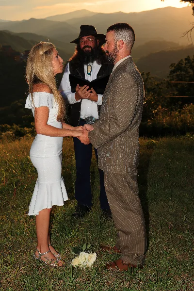 A bearded officiant in a hat and vest stands between a bride and groom holding their hands during an outdoor wedding ceremony with a mountain backdrop.
