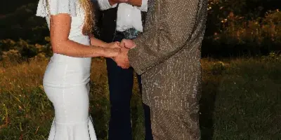 A bearded officiant in a hat and vest stands between a bride and groom holding their hands during an outdoor wedding ceremony with a mountain backdrop.