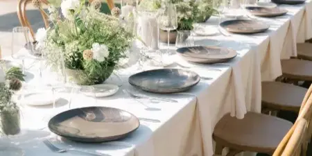 Outdoor wedding reception table featuring white linens, neutral ceramic plates, and lush floral centerpieces with greenery.