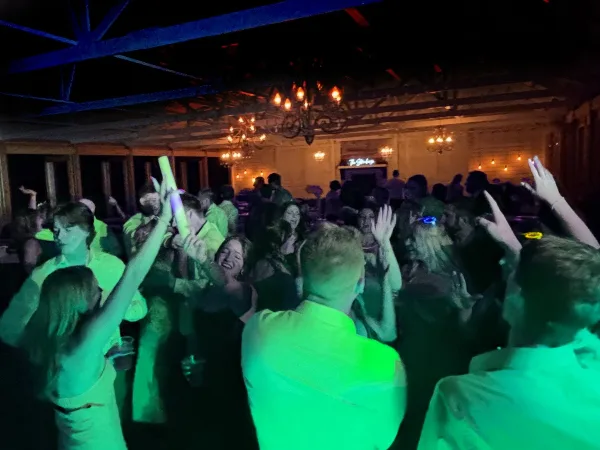 Wedding guests dancing on a crowded dance floor under green and blue event lighting in a venue with chandeliers.
