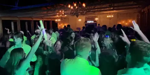Wedding guests dancing on a crowded dance floor under green and blue event lighting in a venue with chandeliers.