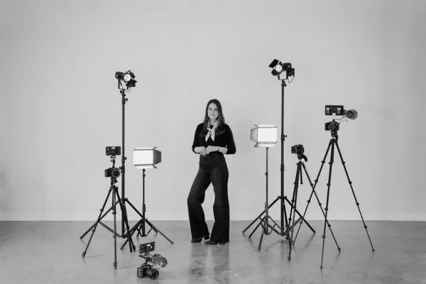 A woman standing in a studio surrounded by professional video cameras, tripods, and lighting equipment.