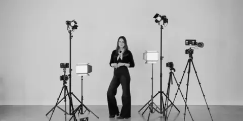 A woman standing in a studio surrounded by professional video cameras, tripods, and lighting equipment.