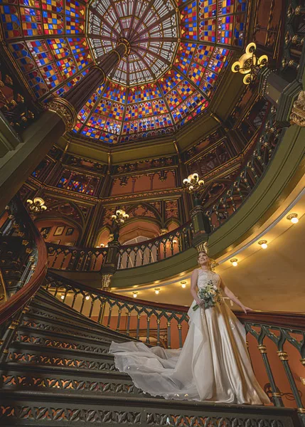 A bride in a white wedding dress stands on a grand staircase with a stained glass ceiling.