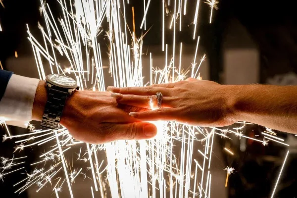 A close-up photograph of a couple's hands holding wedding rings, illuminated by the bright sparks of a sparkler exit.