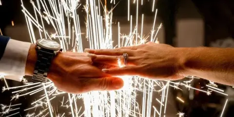 A close-up photograph of a couple's hands holding wedding rings, illuminated by the bright sparks of a sparkler exit.