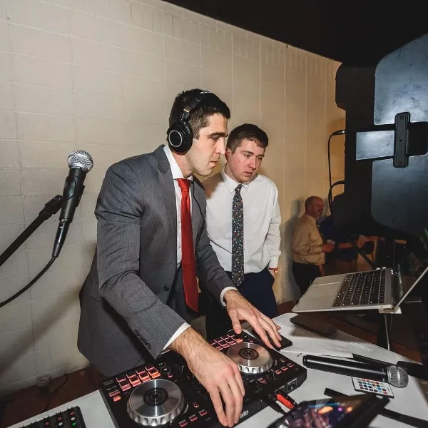 A DJ in a gray suit and red tie mixing on a Pioneer controller with headphones, microphones, and a laptop at an event.