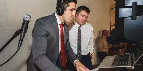 A DJ in a gray suit and red tie mixing on a Pioneer controller with headphones, microphones, and a laptop at an event.