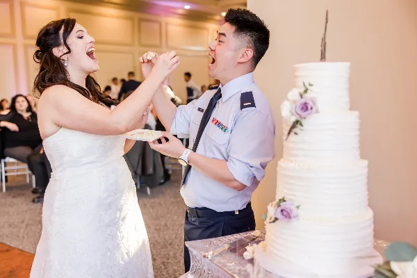 A bride and groom sharing a playful moment with cake at a wedding reception, with a multi-tiered wedding cake in the foreground.