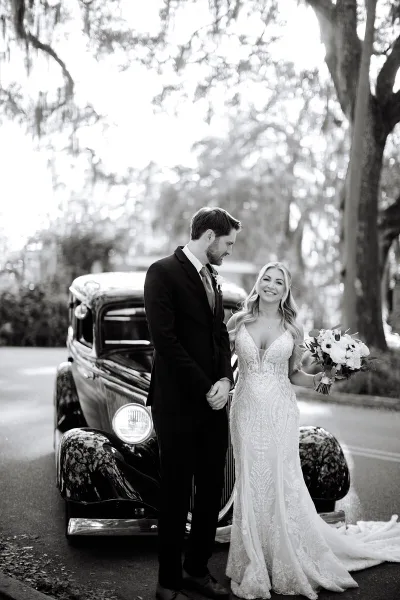 A bride and groom standing together in front of a vintage car, with the bride holding a bouquet of flowers.