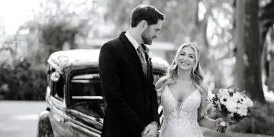 A bride and groom standing together in front of a vintage car, with the bride holding a bouquet of flowers.