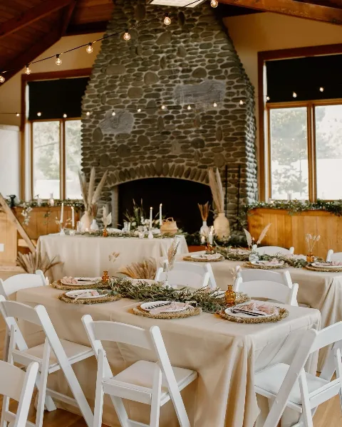 A rustic event venue interior featuring a large stone fireplace and tables set with neutral linens, white chairs, and western boho floral centerpieces.