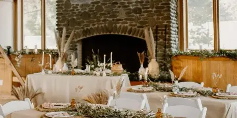 A rustic event venue interior featuring a large stone fireplace and tables set with neutral linens, white chairs, and western boho floral centerpieces.