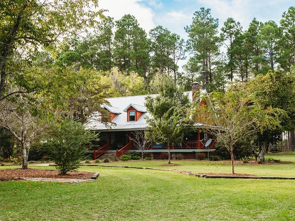 Exterior view of the Main Cabin at Tahoma Plantation, featuring a red roof and porch, surrounded by green lawn and pine trees.