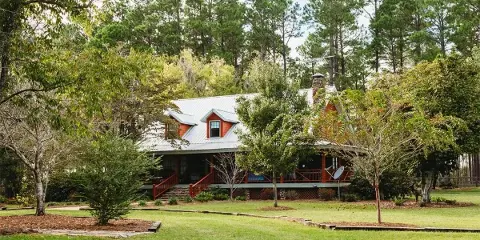 Exterior view of the Main Cabin at Tahoma Plantation, featuring a red roof and porch, surrounded by green lawn and pine trees.