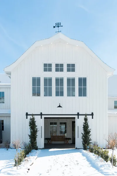 Exterior view of a white modern farmhouse barn with a weathervane and sliding doors in a snowy landscape.