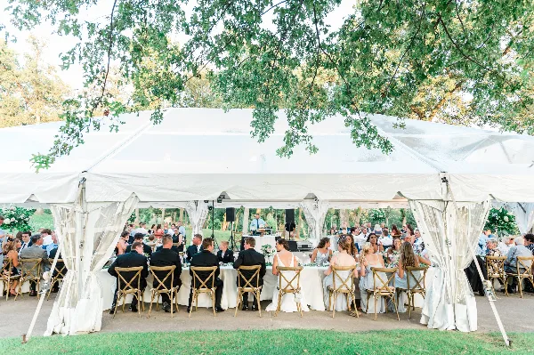 Outdoor wedding reception under a white tent with guests seated at long tables.