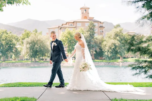 A bride and groom walking hand-in-hand on a paved path near a body of water with a large building in the background.