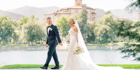 A bride and groom walking hand-in-hand on a paved path near a body of water with a large building in the background.