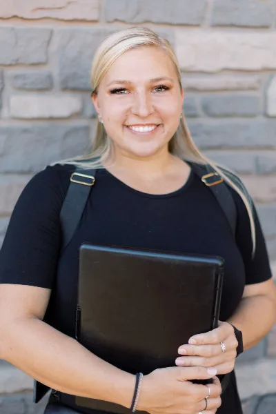 Smiling blonde woman in a black shirt holding a black tablet in front of a stone wall