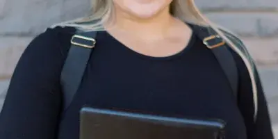 Smiling blonde woman in a black shirt holding a black tablet in front of a stone wall