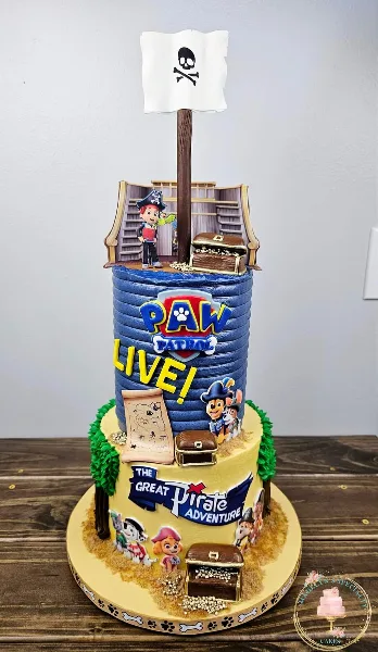 A custom two-tiered cake with a blue top tier and yellow bottom tier, decorated with Paw Patrol characters, pirate ship elements, and a skull and crossbones flag.