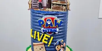 A custom two-tiered cake with a blue top tier and yellow bottom tier, decorated with Paw Patrol characters, pirate ship elements, and a skull and crossbones flag.