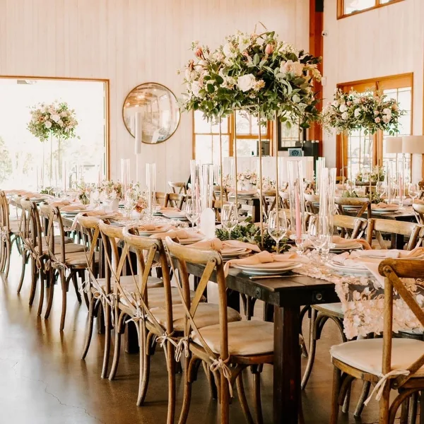 A wedding reception table setting featuring wooden farm tables, cross-back chairs, tall floral centerpieces, and glassware.