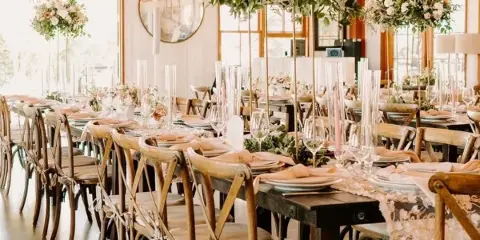 A wedding reception table setting featuring wooden farm tables, cross-back chairs, tall floral centerpieces, and glassware.