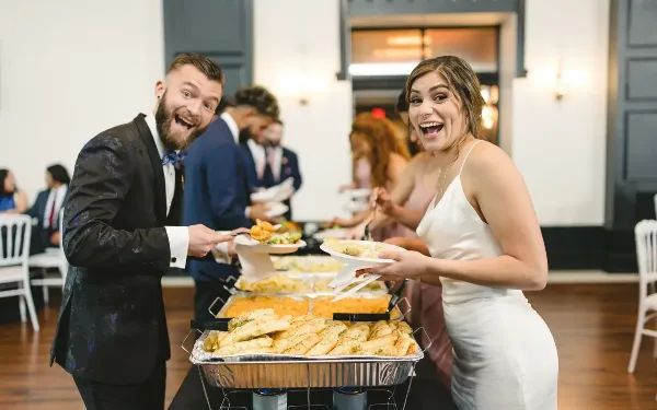A bride and groom serving themselves food from a buffet line with chafing dishes at a wedding reception.