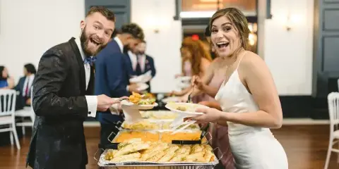 A bride and groom serving themselves food from a buffet line with chafing dishes at a wedding reception.